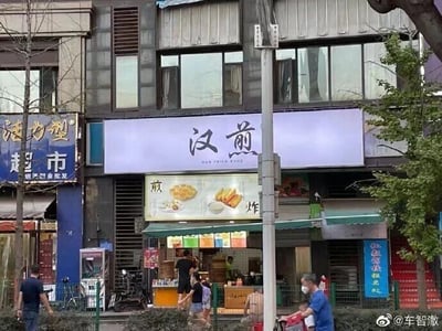 这是一张原始拍摄的城市街景照片，展现了日常的街边商铺与行人活动场景。画面中心是一家煎炸类小吃店，上方的白色招牌醒目地写着“汉煎”二字，下方的灯箱标注“煎炸”并配有食物实拍图，明确店铺经营品类。“汉煎”谐音“汉奸”，是一个趣味谐音梗。店铺左侧是名为“活力型超市”的商铺，右侧也有其他临街店铺。画面中有多位普通行人：有的驻足在小吃店前选购食物，有的在街边步行，还有一位佩戴口罩的市民推着车前行，整体呈现出生活化的城市日常氛围。