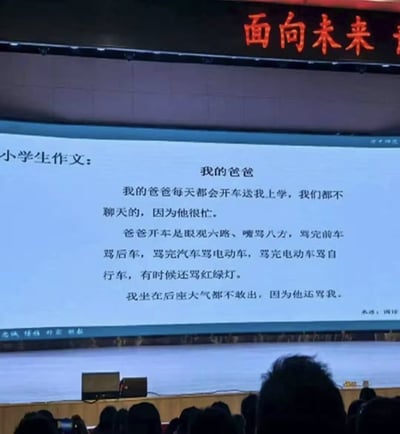 这是一张线下活动现场的实拍照片，拍摄视角位于观众席，画面下方可见多名观众的头顶轮廓，前方的大型展示屏幕上呈现了一篇趣味小学生作文，标题为《我的爸爸》。作文内容以孩子的视角描述爸爸开车送自己上学时的状态：爸爸开车时忙于咒骂路上的前车、后车、各类交通工具甚至红绿灯，全程没空和孩子聊天，孩子坐在后座不敢出声生怕也被爸爸骂，内容夸张诙谐，生动刻画了路怒司机的日常状态，屏幕顶部还能看到部分红色的“面向未来”字样。