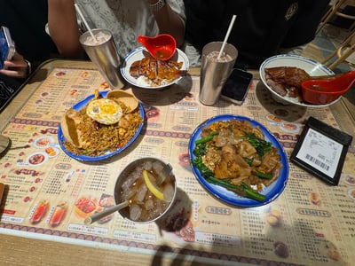 图片展示了一张摆满港式美食的餐桌，桌上有两份烧鸭饭、一盘炒饭（上面有一个煎蛋）、一盘炒河粉（配有青菜），以及三杯冰镇饮品和一碗柠檬饮品。桌子表面铺着印有各种菜品图片和文字的餐垫，旁边可以看到部分用餐人的手臂和手机。整体场景呈现出在香港餐厅用餐的氛围。