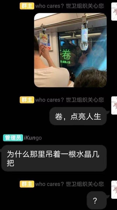 这是一段即时通讯群的聊天记录截图，群主多次发送固定签名消息并分享了一张地铁内的实拍照片：地铁门屏幕上显示着“卷，点亮人生”的标语，旁边还有一个透明的地铁吊环。群成员先重复了标语内容，随后管理员看错了透明吊环的形状，发言“为什么那里吊着一根水晶几把”，引发搞笑效果，最后群成员发出问号表示疑惑。
笑点在于管理员对地铁透明吊环的视觉误解，将普通的吊环看成了不雅的形状，制造出沙雕的反差感。