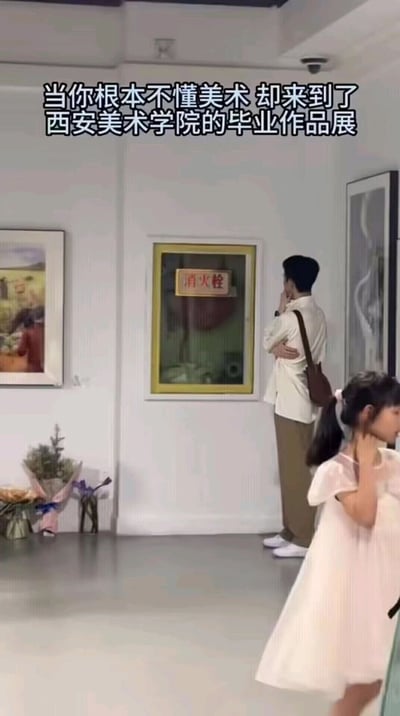 这是一张拍摄于西安美术学院毕业作品展现场的实拍图，画面顶部配有文字“当你根本不懂美术 却来到了西安美术学院的毕业作品展”。画面中，一名身着浅色上衣、背着斜挎包的男子站在墙面上的消火栓前，摆出一手托着下巴、一手抱臂的姿态，仿佛在认真品鉴这件“艺术作品”，但实际上他错把消火栓当成了展出的毕业作品。画面左侧挂有正规的参展画作，右下角有一名小女孩正从旁走过，整个场景通过男子的乌龙行为，生动且搞笑地展现出不懂美术的人置身艺术展时的茫然与尴尬，充满反差感与趣味性。