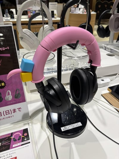 电子产品商店展示柜中，一款设计独特的耳机成为焦点。这款黑色耳机配备了造型夸张的粉色头梁，其弯曲形态酷似动物的脖颈或犄角，末端还装饰有蓝色和黄色的立方体元素，整体设计充满趣味性和创意感。背景中可以看到其他款式的耳机和带有日文文字的宣传海报，显示这是在日本的一家电子产品零售店。