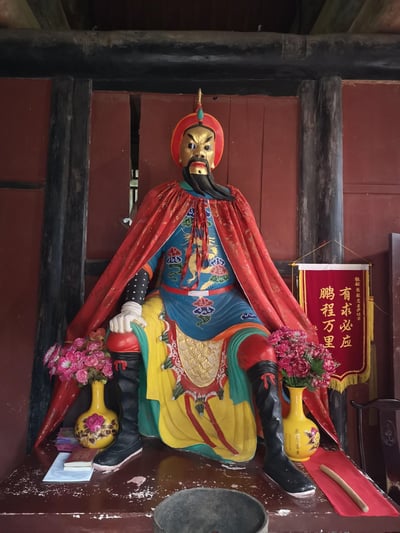 这是一张室内供奉场景的实拍照片，主体是一尊彩绘的明末农民起义领袖张献忠的塑像：塑像面容涂金、神态威严，身着蓝底绣龙袍服，外披红色织金披风，端坐于供台之上。供桌上摆放着插有粉色仿真花的黄色花瓶、经书、香炉等供奉物品，塑像右侧悬挂着一面红底金字的锦旗，上书「敬献张献忠菩萨位前 有求必应 鹏程万里」，呈现出民间将历史人物作为信仰对象供奉的场景。