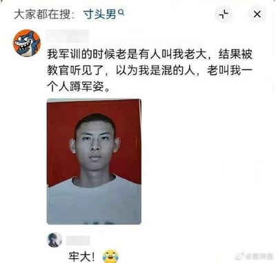 这是一张社交媒体帖子截图，顶部显示平台搜索热点为“寸头男”。发帖用户头像是卡通鲨鱼形象，配文称自己军训时总被同学喊老大，被教官听见后误以为他是混社会的人，经常单独罚他蹲军姿，配文下方附上了该用户的寸头红底证件照，照片里的男生外形硬朗气质偏严肃。下方评论区有网友回复“牢大！”搭配搞怪表情，玩谐音梗，“牢大”谐音“老大”，调侃该男生的外形看起来很有社会感，笑点十足。