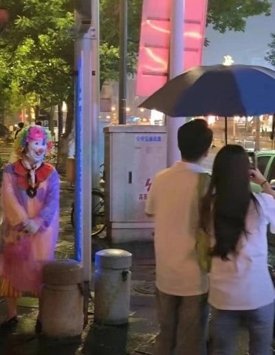 雨夜的城市街头，地面因雨水变得湿滑反光，一对情侣正共撑一把黑伞背对镜头，似乎在专注于眼前的事物或拍照。而在他们的侧后方，柱子旁站着一位身着色彩渐变的粉紫色长外套、戴着诡异笑脸面具的小丑，双手交握放在身前，安静地注视着这对情侣，整体氛围带着强烈的惊悚感，仿佛下一秒就会发生让人不安的事，给人一种背后发凉的诡异体验。