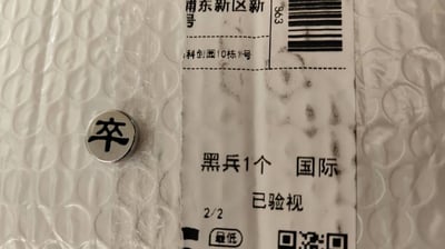 图片展示了一张带有气泡膜的快递包裹局部，上面贴有快递标签。标签上包含地址信息“浦东新区新号 科创园10栋1号”、物品描述“黑兵1个 国际”、验视信息“已验视”、编号“2/2”及“最低”字样，右侧有条形码和二维码。在标签左侧的气泡膜上，放置着一枚金属材质的象棋棋子，棋子表面刻有黑色的“卒”字（中国象棋中的兵卒）。整体为真实拍摄的日常场景，无明显双关、谐音梗或暗示内容。