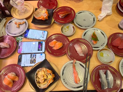 图片展示了Sushiro寿司店的餐桌场景，木质桌面上摆满了各色寿司和料理。主要有红色和灰色两种带有“スシロー”字样的盘子，盛放着三文鱼寿司、蟹腿、鱼肉寿司等，还有黑色方形盘子装着烤蟹壳。桌上有三杯透明饮料、三包手机（屏幕亮着，显示疑似菜单或社交媒体内容），以及少量用餐后的纸巾。整体呈现出丰盛的日式料理用餐氛围，充满食欲感。