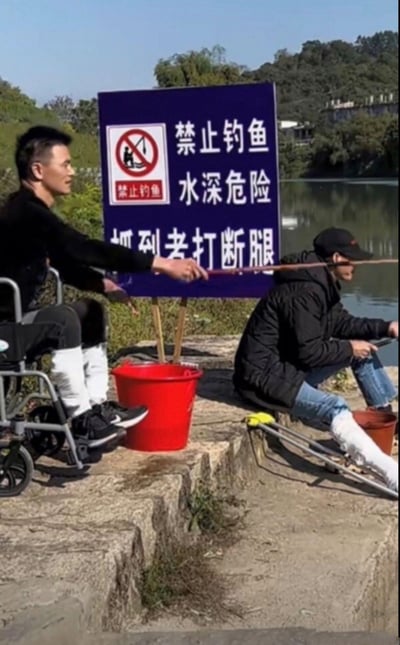 在一处绿植环绕的水边，立着一块深蓝色警示标语牌，牌面标注“禁止钓鱼 水深危险 抓到者打断腿”，还有标准的禁止钓鱼标识图案。标语牌前有两位男子，一位坐在轮椅上，双腿佩戴白色护具，面带笑意手持钓鱼竿；另一位同样腿戴护具，坐在简易的移动装置上专注钓鱼。两人的状态和标语里的夸张惩罚“打断腿”形成强烈反差，用黑色幽默的方式玩梗，仿佛在暗示他们就是违反禁令被“惩罚”的人，却依然坚持钓鱼，充满戏谑感。