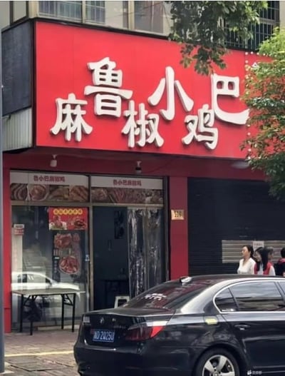 图片展示了一家位于城市街道旁的餐馆，红色招牌十分显眼，上面用白色大字写着“麻鲁小椒鸡”。招牌下方是玻璃门，门上贴有食物图片和宣传海报。店门前停放着一辆黑色轿车，路边有几位行人经过，周围有绿树点缀，整体呈现出日常街头的景象。