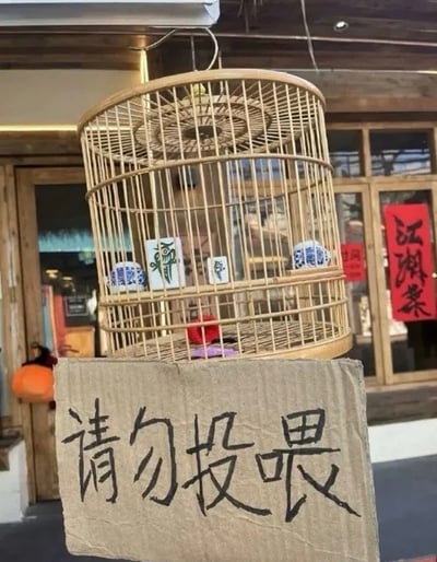 这是一张线下场景实拍图，画面主体是悬挂在店铺外的竹制圆形鸟笼，鸟笼内摆放两块麻将牌，拼出“穷鬼”二字，还搭配了两个传统蓝白瓷鸟食罐；鸟笼前方垂挂一块粗麻布，麻布上用黑色毛笔书写了“请勿投喂”四个大字；背景是带木质门窗的店铺门面，右侧玻璃可见红底黑字的“江湖菜”字样，属于商家利用当代年轻人穷鬼自嘲的网络梗设置的趣味打卡布置。