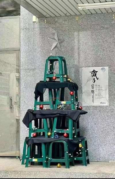 室内或半户外场景中，多层绿色塑料凳堆叠成金字塔形创意圣诞树，凳间用黑色布料遮盖缝隙，外层缠绕白色串灯并悬挂红金球形装饰。背景为灰色大理石墙面，上方有星形装饰，右侧贴有“歲月留聲”字样及“12.13-14”日期的海报，展现低成本环保的节日装饰巧思，充满生活气息。
