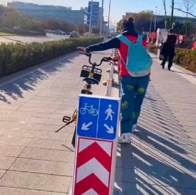 这是一张街头拍摄的趣味视觉错位照片：画面中的行人下半身与前方的交通分道指示牌（左侧指示自行车道、右侧指示行人道）的自行车图案、旁边的自行车把手恰好重合，从拍摄视角看去，仿佛这个人正骑着指示牌上的“自行车”，制造出荒诞又搞笑的视觉错觉，没有双关或谐音梗。