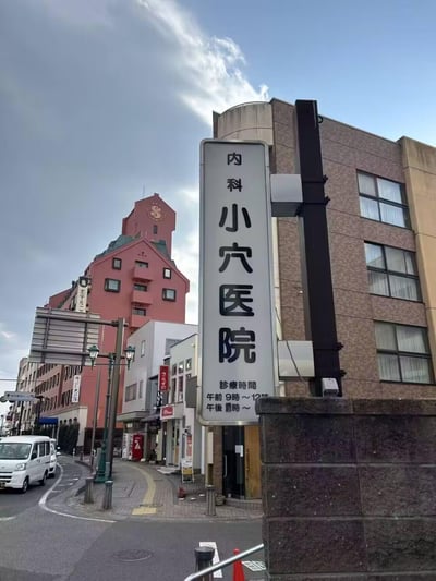 这是一张在日本街头拍摄的日常街景照片，画面视觉中心是一块竖版的白色医院招牌，上面用日文写着“内科 小穴医院”，下方标注了诊疗时间（午前9时～12时，午後时间部分被遮挡）。招牌的字面意思容易让人产生趣味联想。背景是具有日式风格的多层建筑，其中一栋造型独特的红色尖顶建筑十分醒目，街道上停着白色车辆，还有临街的小型店铺，整体呈现出日本居民区附近的日常城市风貌。