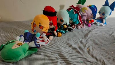 照片中展示了一排色彩丰富的动漫角色plush玩具，整齐摆放在铺有蓝白条纹床单的床上。这些玩具造型各异，拥有橙色、红色、白色、青色、粉色等多种发色，部分佩戴帽子、蝴蝶结等装饰，服装细节丰富，背景为简洁的浅色墙面，整体呈现出温馨可爱的收藏场景。