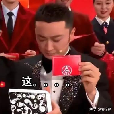 图中展示了一场晚会或大型综艺节目舞台，穿着礼服的人物手持红色卡片和魔术道具，卡片上印有类似和平标志的符号。图片底部大字写着“这……”表达困惑或无语氛围，非常适合用来形容尴尬、翻车或意外出现时的情绪。画面幽默，带有错愕和想吐槽的气息，现场感十足。未发现明显双关或谐音梗，但整体隐含“魔术失败”等反差喜感。