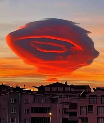 这张图片呈现了黄昏时分的城市上空景致，画面下方是几栋错落的多层居民楼，楼体外观是常见的城市住宅样式，一盏路灯已经点亮，传递出日暮的氛围感。天空中悬浮着一块形态极为罕见的荚状云，在夕阳的渲染下，云朵的边缘被晕染出炽热的橙红色，云朵内部的层次恰好构成了类似人类眼睛的轮廓，宛如天空睁开了一只巨大的眼眸，视觉效果极具冲击力，给人带来奇幻又震撼的观感，让人惊叹于自然气象的神奇造物。
