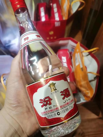 一只手手持一瓶汾酒，瓶身为透明玻璃材质，标签为红白配色，印有“汾酒”字样及山西杏花村汾酒厂股份有限公司出品等信息。背景中可见红色和橙色的包装礼品袋，整体场景像是在展示或准备赠送的白酒产品。图片清晰展示了汾酒的外观细节和包装环境。