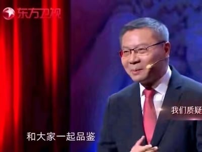 图片中是一位身着黑色西装、白色衬衫搭配红色领带的男子，他戴着眼镜，头戴麦克风，站在舞台上。背景为红色窗帘和紫色渐变幕布，左上角有“东方卫视”的台标。男子面带温和微笑，似乎正在进行发言，画面底部出现字幕“和大家一起品鉴”与“我们质疑”，整体场景呈现出电视节目中的演讲或评论环节。