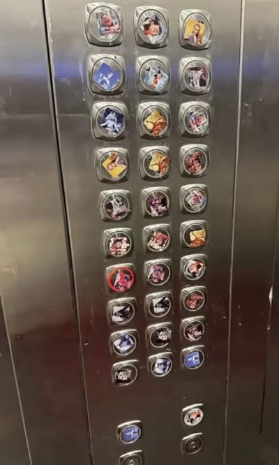 这是一张竖幅实拍照片，展示了一块不锈钢材质的电梯楼层按键面板。面板上的所有楼层按键几乎都被人贴上了圆形的自定义贴纸，贴纸内容各不相同，包含真人表情包、卡通动漫人物、网红梗图等，完全覆盖了原本按钮上的楼层数字，只剩最底部两个按键未被贴纸遮挡，属于网友在公共电梯里的恶搞行为，整体内容滑稽有趣。