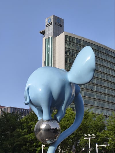 图片展示了一座大型蓝色大象雕塑，雕塑整体呈流线型，大象后腿站立在一个光滑的银色球体上，长长的鼻子自然下垂。背景是一栋高层建筑，顶部有明显的“CCB”标志，推测为中国建设银行的办公楼。雕塑位于前景，周围有绿色树木点缀，天空为晴朗的蓝色，整体画面现代感与艺术感兼具，展现了城市公共空间中的艺术装置与金融建筑的结合。