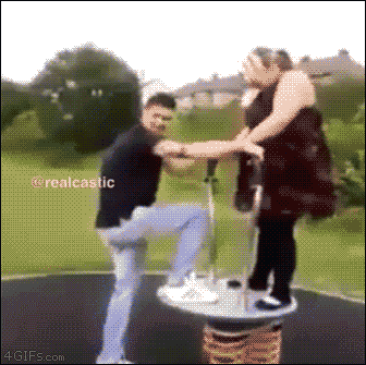 这是一个动态图（GIF），画面背景是户外草坪区域，能看到远处的房屋和树木。画面中有一个弹簧弹跳装置，一名穿着黑色上衣、浅蓝色裤子的男子正试图登上该装置，他拉着站在装置上的黑衣女子的手，结果装置失去平衡，女子身体剧烈晃动，面露惊慌试图维持平衡，男子也没能顺利站稳，身体跟着失衡，整个过程充满滑稽感，是日常户外玩耍时的搞笑失误场景。