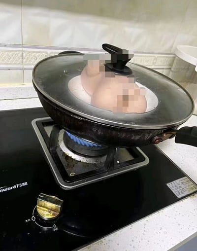 一张厨房场景照片，燃气灶上的炒锅中透过玻璃盖隐约可见疑似人体部位的模糊物体，营造出诡异惊悚的视觉效果。实际上这可能是利用角度和物品摆放形成的视觉错觉或刻意摆拍的恶作剧图片。