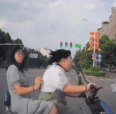 两位女生骑着电动车在城市十字路口经过，画面抓拍到一人坐驾驶座，一人坐后座，气氛轻松，有生活的烟火气。后方背景有红灯笼灯饰，正值绿灯通行。虽是抓拍，整体充满青春活力感。适合用来表达出门游玩、姐妹出行、赶时间上学等场景。

画面无明显双关、谐音梗，也未见暗示成分，属于生活抓拍类型。