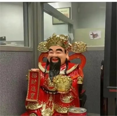 这是一个在办公室隔间拍摄的趣味合成梗图，将人物的脸替换到了财神爷的财神像上，仿佛“真人版财神”坐镇办公区。财神塑像手中托着元宝，另一只手拿着写有“招财进宝”的牌子。整体场景很有喜感，类似cosplay/现实融梗，体现了人们对办公室财运和好运的美好期望。

没有谐音梗，但通过“真人版财神”形成视觉冲击，有种幽默的‘我要做财神’的暗示。