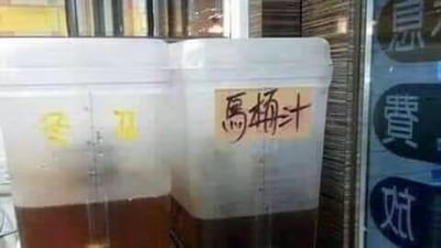 图片展示了左右并排的两个透明塑料桶，桶内装有深棕色的液体，看起来是对外售卖的桶装饮品。右侧桶身用马克笔写着‘馬桶汁’（马桶汁）的字样，左侧桶身有模糊的黄色手写文字。这种用极具违和感、让人不适的词汇来命名饮品的方式，制造出强烈的反差感，显得十分搞笑离谱，很容易引发网友的调侃。