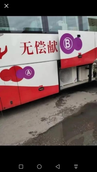 图片展示了一辆无偿献血车的侧面。车身主体为白色，配有红色装饰条纹，车身上有醒目的红色中文文字“无偿献血”。在文字旁边和下方，有两个紫色圆形标识，分别标有“A”和“B”字样，代表A型血和B型血。车辆停放在室外，地面有一些水渍和污渍。