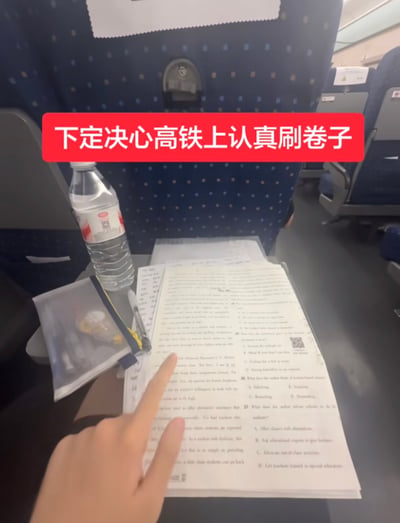 这张实拍图的拍摄场景为高铁车厢内部，画面下方是拍摄者伸出的手，手指正指着小桌板上摊开的英语练习题试卷，桌板上还摆放着瓶装矿泉水、透明文具袋和笔，背景是高铁典型的深蓝色带浅色圆点的座椅靠背。图片上方叠加了红底白字的醒目文字“下定决心高铁上认真刷卷子”，搭配原标题传递出网友想要在出行途中好好学习的初衷，暗含后续大概率无法坚持、调侃自己是享福命的自嘲趣味，很容易引发有类似经历网友的共鸣。
