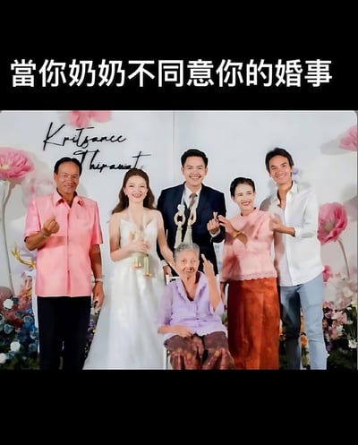 一张婚礼合影照片，画面中有六位穿着正式服装的人，包括一对新婚夫妇和四位长辈。所有人都面带微笑，站在装饰有粉色花朵的背景前。然而，坐在中间的奶奶却做出了一个明显的不同意手势，与整体欢乐的氛围形成鲜明对比。图片上方有文字“當你奶奶不同意你的婚事”，通过奶奶的手势和文字结合，形成了幽默的反差效果。