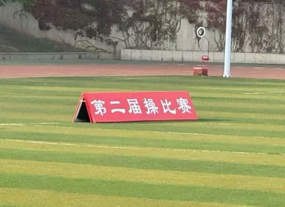 一张在操场草坪上拍摄的红色指示牌照片，上面写着"第二届操比赛"白色大字。背景可见跑道、石墙和运动场地设施，整体场景像是学校或体育场馆的室外运动场。