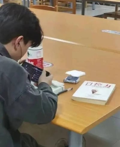 一名男子坐在食堂或自习室的木质餐桌前，戴着耳机低头专注看手机。桌上放着一本名为《自控力》的书、一个红白相间的杯子、一包纸巾和一盒香烟，形成了书本主题与实际行为的有趣反差——本应提升自控力，却在沉迷手机。
