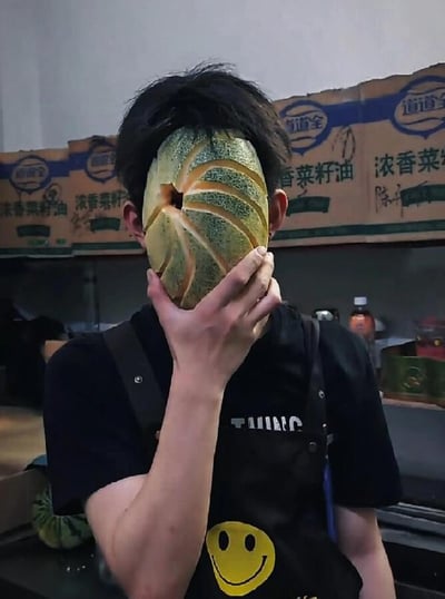 一个人站在厨房环境中，用一个切开的哈密瓜当作面具遮住了整个脸部，只露出眼睛的位置。他穿着一件黑色的T恤和带有黄色笑脸图案的黑色围裙，背景中可以看到堆放的食用油纸箱，上面有"浓香菜籽油"等字样。整个场景看起来像是在食品加工或厨房工作场所拍摄的趣味照片。
