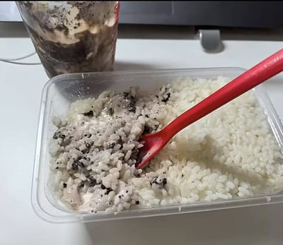 图片展示了一份白米饭被端放在塑料餐盒中，其中一部分米饭上覆盖着奥利奥冰淇淋般的混合物，旁边还放着一只红色塑料勺。画面后方还有一个装有奥利奥冰淇淋或麦旋风的纸杯。这种将甜品和正餐主食混合的吃法看起来十分离谱，引发强烈违和感和网友的吐槽。此类图片常用于吐槽学生党、单身狗或外卖党生活的节俭、无奈或离谱操作，也可作为“食物黑暗料理”的典型例子出现。

该图充满搞笑、离谱和一点恶趣味的氛围，暗含对现实压力和无奈生活的自嘲，属于典型的“黑暗料理”梗。

如有需要，这类组合可以作为对“你能接受这种吃法吗？”、“有勇气挑战吗？”等网络讨论的示例。