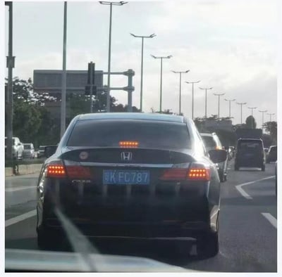 这是一张在城市道路车流中拍摄的实拍图，画面主体是一辆黑色广汽本田轿车的车尾，车辆的左右刹车灯亮起，加上后窗中间的一排红色小灯，三者组合起来恰好形成了类似带有无奈、囧态的人脸表情，十分搞笑。车牌为粤K FC787，背景中可以看到城市道路的路灯、其他行驶的车辆以及道路旁的绿植，整体是日常驾车途中偶然捕捉到的趣味巧合场景，没有刻意的后期修改，充满生活中的小趣味。