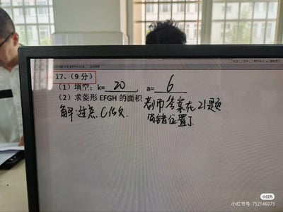 这是一张翻拍电脑屏幕的照片，屏幕显示的是一份数学试卷的第17题，该题总分9分，其中（1）的填空已经填写了答案k=20、a=6，（2）要求求解菱形EFGH的面积，解题步骤仅写了开头“解:过点C作...”，旁边用手写字体标注了“老师答案在21题写错位置了”，透露了老师批改试卷时的小失误。画面背景能看到两位真实人物，似乎处于教室或办公场所，整体是校园里的搞笑乌龙场景，充满日常感。