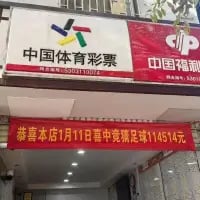 这是一张实拍的线下门店照片，画面中相邻开设着中国体育彩票与中国福利彩票的门店，门店外悬挂着一条醒目的红色横幅，上面印有文字“恭喜本店1月11日喜中竞猜足球114514元”，直观展示了该体育彩票门店中出大额竞彩足球奖金的喜讯，整体画面构图清晰，信息明确。