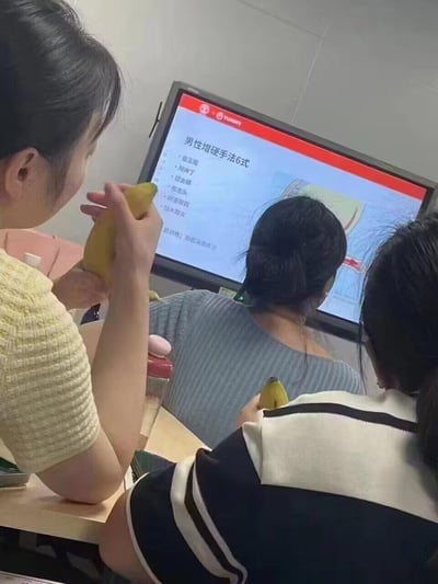这是一场线下性健康技能培训的现场：几位女性学员坐在桌前，前方的教学屏幕上展示着「男性增硬手法6式」的详细内容，包含盘龙圈、锁神丁、搓金棒、捏龙头、阴茎颈段、钻木取火6种手法，以及「肌训练：勃起消退练习」的补充内容。画面中的学员手持香蕉，将其作为模拟道具，实操练习教学的手法，整体场景是针对男性健康护理的职业技能培训，用香蕉作为模拟物的设定带有一定的反差感，显得较为猎奇。