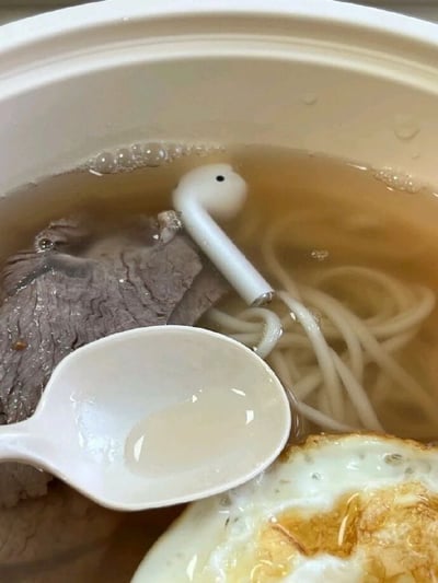这是一张实拍的生活类搞笑图片，画面主体是盛放在白色餐盒里的汤米粉，汤中还有酱色的牛肉片、半块煎蛋以及一个白色塑料汤勺。一只白色的AirPods无线耳机不慎掉进汤里，耳机的长杆部分和旁边白色的米粉颜色、粗细相近，视觉上形成了耳机正在吸食米粉的错位效果，十分有趣。