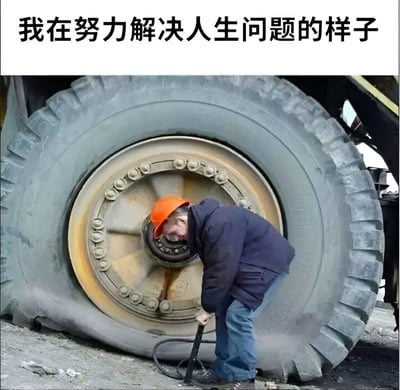 图片上方配有文字'我在努力解决人生问题的样子'，下方是一名戴着橙色安全帽、身穿深色外套的男子，正弯腰使用工具对着一个巨大的车辆轮胎进行操作。通过男子面对巨大轮胎显得渺小却仍在努力的场景，幽默自嘲地比喻人们在面对人生难题时看似努力却可能收效甚微的状态。