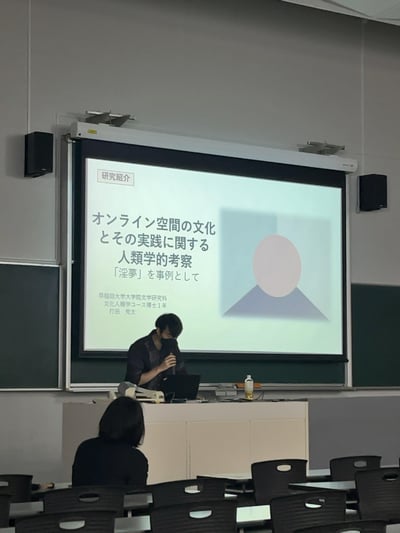 这是在早稻田大学教室的学术发表场景，文学研究科文化人类学课程的博士一年级学生打田秀太，正在进行一场关于网络亚文化的人类学研究汇报。讲台后的投影屏幕清晰展示了研究标题《关于网络空间的文化与实践的人类学考察——以「淫梦」为案例》，以及发表者的院校、年级和姓名信息。讲台上的学生手持话筒，低头似乎在准备发言或整理思路，教室里还有其他听众在场。
注：「淫梦」是日本NICONICO平台诞生的经典网络亚文化迷因，将其作为学术研究案例，是亚文化进入学术视野的有趣跨界表现。
