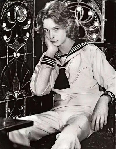 A black and white vintage photograph featuring a young person with curly hair wearing a sailor-style outfit. The subject is seated in front of an ornate metal background with circular decorative elements. They have their right hand resting on their cheek and are looking directly at the camera with a serious expression. The sailor uniform includes a collared shirt with striped cuffs and a necktie, paired with matching pants.