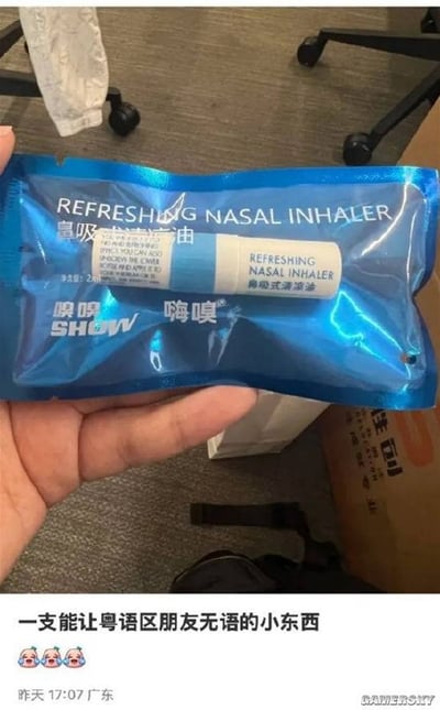 图片中一只手握着一包蓝色包装的鼻吸式清凉油，包装上印有英文“REFRESHING NASAL INHALER”和中文“鼻吸式清凉油”，品牌为“嗅嗅”和“嗨嗅（SHOW）”。背景可见办公室的灰色地毯、黑色办公椅轮子和部分纸箱。图片下方配有文字“一支能让粤语区朋友无语的小东西”，因“嗅嗅”和“嗨嗅”在粤语中发音可能接近不雅词汇，从而产生幽默效果，让粤语使用者感到尴尬又好笑。