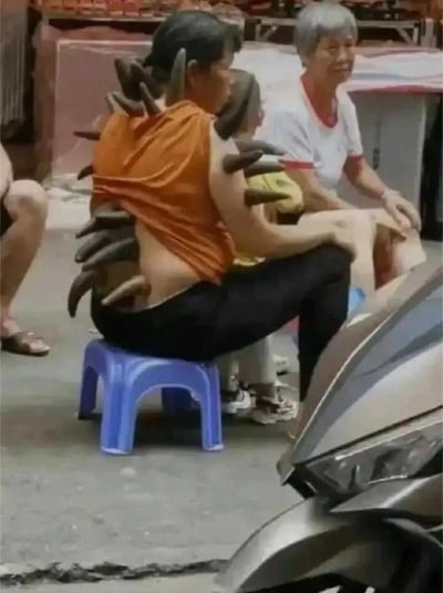 这是一张街头生活化场景的实拍图，画面核心是有趣的视觉错位效果：一位身着橙色无袖上衣的女士坐在蓝色塑料矮凳上，从拍摄视角看去，她的背部仿佛“长”出了一排深色尖刺，营造出类似怪兽的奇特视觉效果。实际上，这些尖刺是女士身后的孩子所佩戴的恐龙造型装饰（帽子或背包），因位置巧合与女士背部重叠，才制造出这个搞笑的错觉。画面右侧可见一辆银色电动车的车头部分，背景中还有其他坐着的路人，整体场景充满生活气息，而这个偶然的错位让普通日常变得趣味十足。