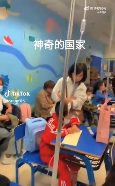 这是一段实拍视频的截图，画面呈现了一个类似教室的空间：一名身着红色校服的小学生坐在蓝色课桌前写作业，手臂上连接着输液的吊瓶，输液杆立在课桌旁。周围还有其他孩子和陪同的家长，环境的墙面有卡通风格装饰，布置和教室高度相似。图中配文“神奇的国家”，点出这种让生病的孩子在类教室环境中一边接受输液治疗一边学习的特殊场景，传递出一种心酸又无奈的氛围。