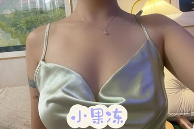 图片中展示了一位穿着浅绿色缎面吊带的女性，领口呈深V设计，凸显优雅气质。她佩戴着一条精致的项链，吊坠小巧别致。左臂上有一处纹身，增添了个性色彩。背景中可以看到一幅绿色植物画作、一盏黄色台灯和米色沙发，营造出温馨的室内氛围。图片底部有一个云朵形状的文字框，内有蓝色字体的“小果冻”字样，可能是对穿着者的昵称或某种可爱的形容。