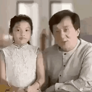这是一段搞笑动态GIF，内容是成龙在画面中先是抬手做出要拍打身旁小女孩头部的动作，小女孩露出无奈又略带紧张的表情，结果成龙的手最后落在了自己的头上，随后他转头对着镜头露出笑容，整个过程通过动作的反差制造出喜剧效果，没有双关、谐音梗等内容。