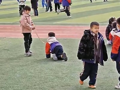 这是小学操场的课间场景，画面里一个穿着粉色外套的小女孩正拉着绳子，绳子套在一个趴在塑胶跑道旁草坪上的小男孩身上，两人像是在玩模仿“拉宠物”的游戏；周围还有其他穿着校服的小朋友，有的在走动，有的在一旁玩耍，整个场景充满了小学生的天真趣味，是孩童间随性又欢乐的日常互动。